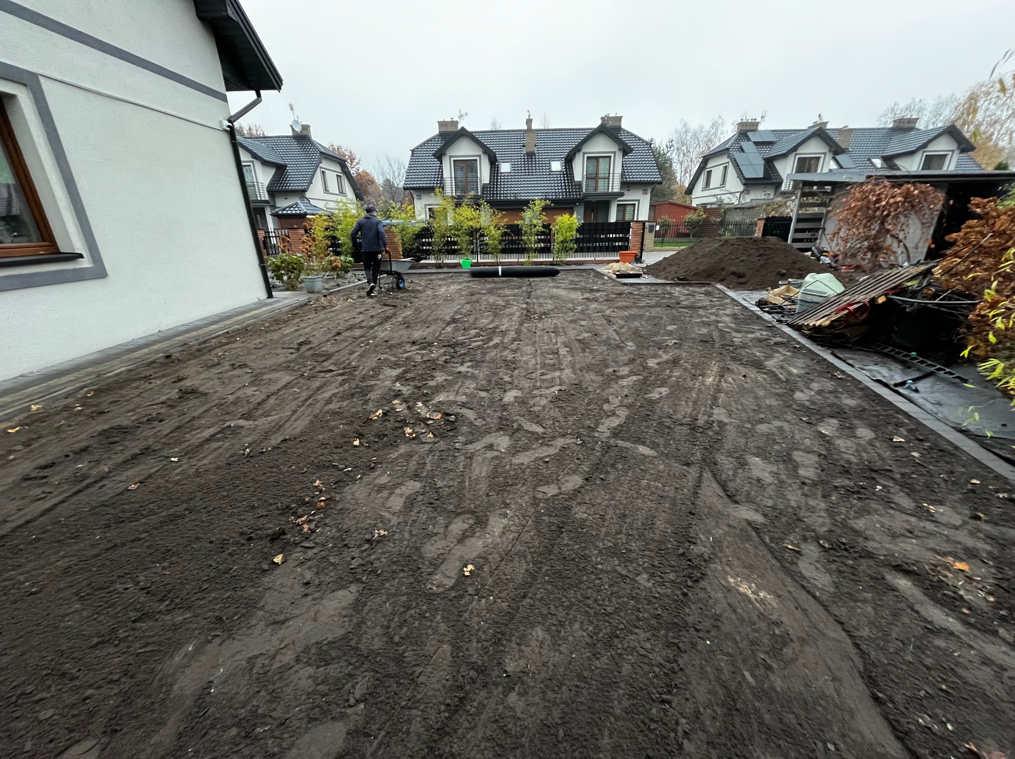 Przygotowanie terenu pod trawnik: wyrównana ziemia, widoczne rolki trawy, osoba z taczką. W tle domy z ciemnymi dachami i ogrodzenie.