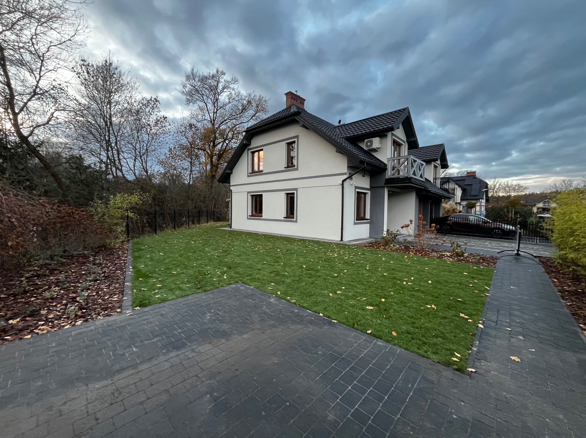 Nowoczesny dom z zadbanym trawnikiem i kostką brukową. Widok na elewację, regularnie skoszony trawnik z opadłymi liśćmi i fragment podjazdu z ciemnej kostki.