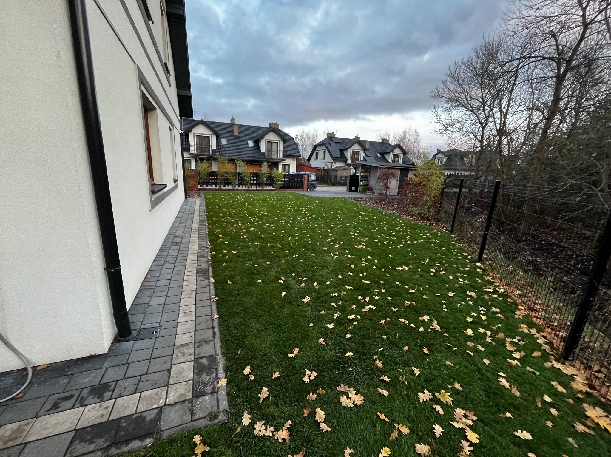 Starannie skoszony trawnik z opadłymi liśćmi dębu, przylegający do białego domu z ciemną kostką brukową. W tle domy z ciemnymi dachami pod pochmurnym niebem.