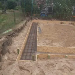 Przygotowanie podłoża pod fundament: szalunki z płyty OSB i zbrojenie stalowe w wykopie. Widoczne drewniane podpory i piasek. Prace ziemne w toku.
