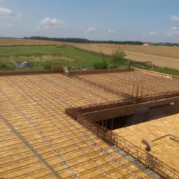 Zbrojenie płyty fundamentowej na deskowaniu z płyty OSB, widok z góry na rozległe pole i zabudowania w tle. Przygotowanie do wylewki.