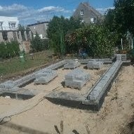 Zbrojenie fundamentów pod budynek mieszkalny. Betonowe bloczki, stalowe pręty, piasek. Widok z góry, w tle dom jednorodzinny i zieleń.