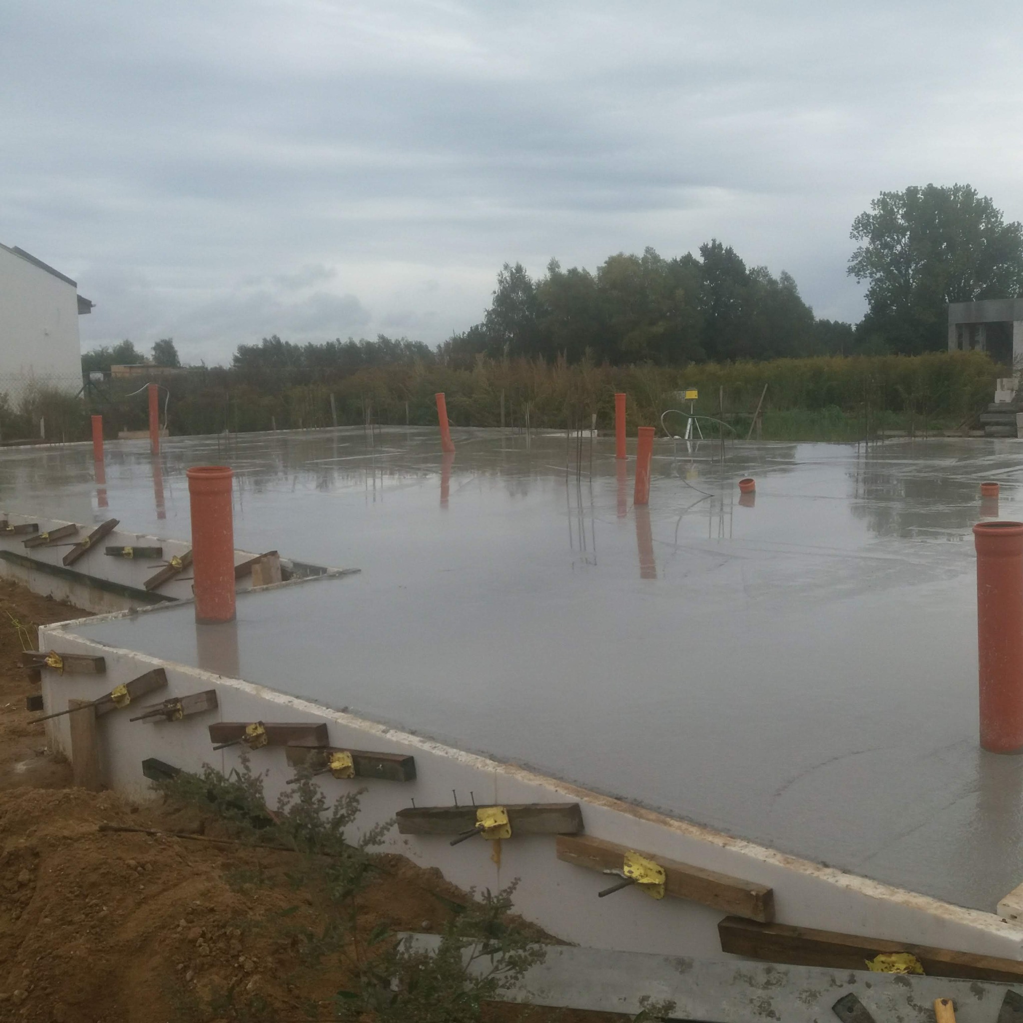 Świeża wylewka fundamentowa z wystającymi rurami kanalizacyjnymi, wykonana na zewnątrz budynku w pochmurny dzień. Widoczne szalunki i elementy konstrukcyjne.