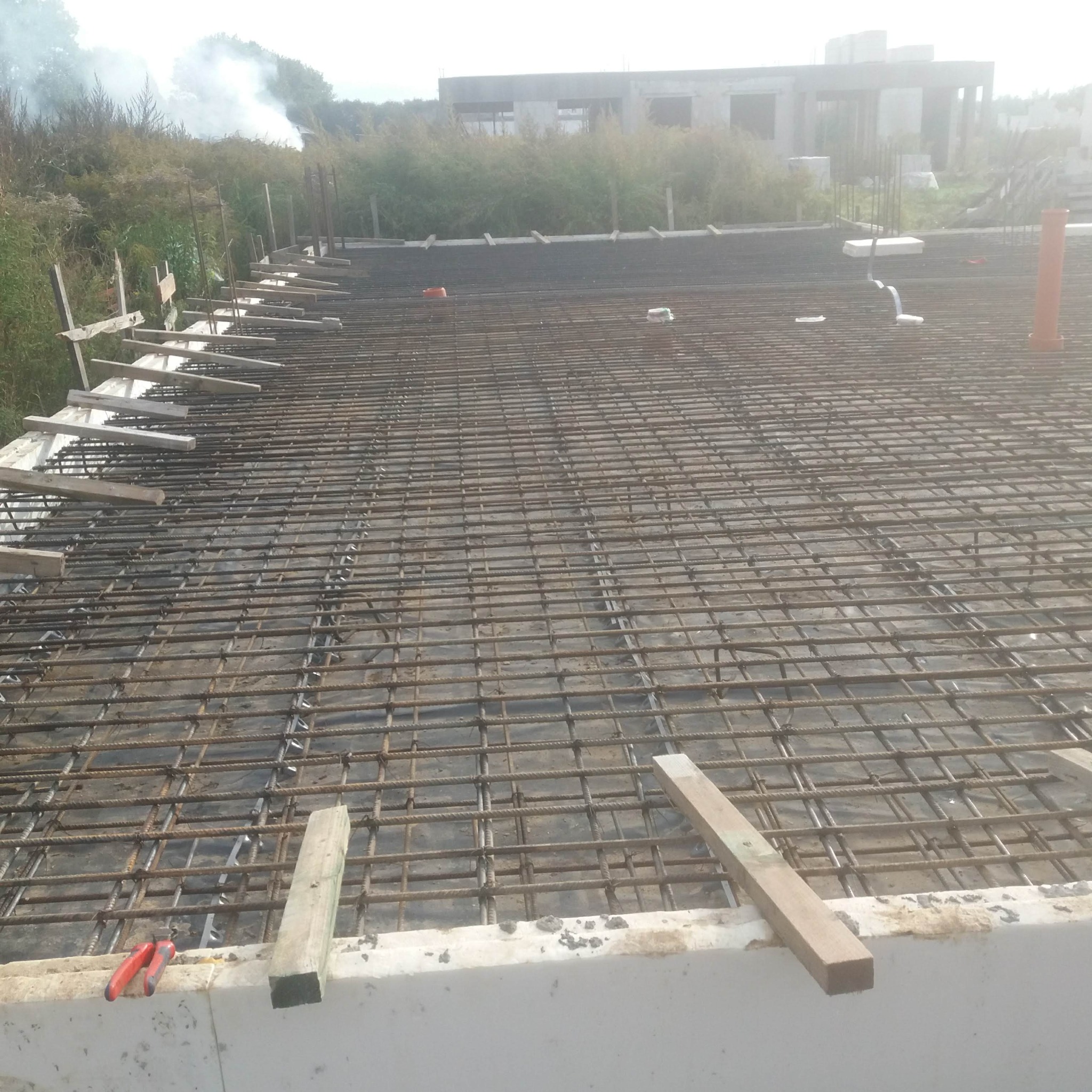 Zbrojenie stropu na budowie, widoczne pręty stalowe, drewniane podpory i fragment konstrukcji budynku w tle. Na pierwszym planie leżą obcęgi.