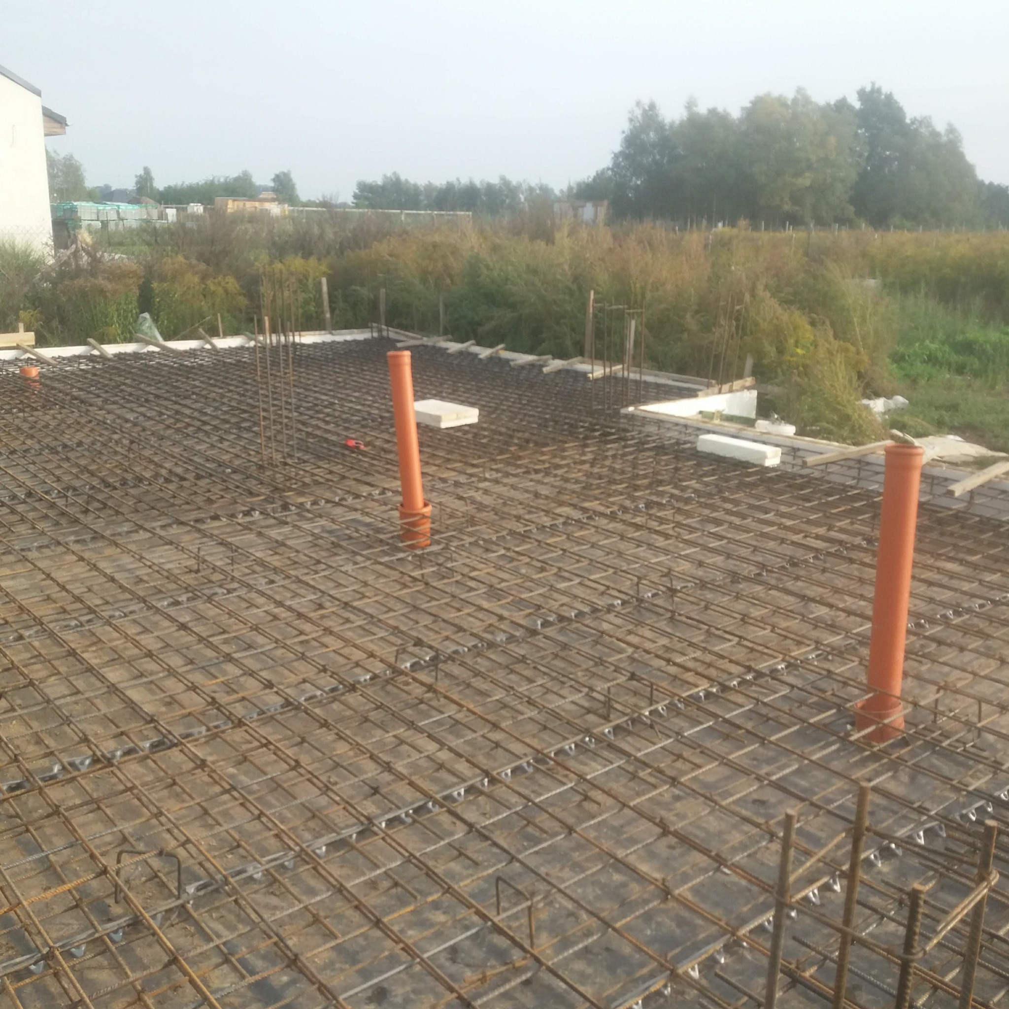 Zbrojenie płyty fundamentowej z wystającymi rurami kanalizacyjnymi, widok z góry. Na drugim planie zieleń i budynek w oddali, pochmurna pogoda.