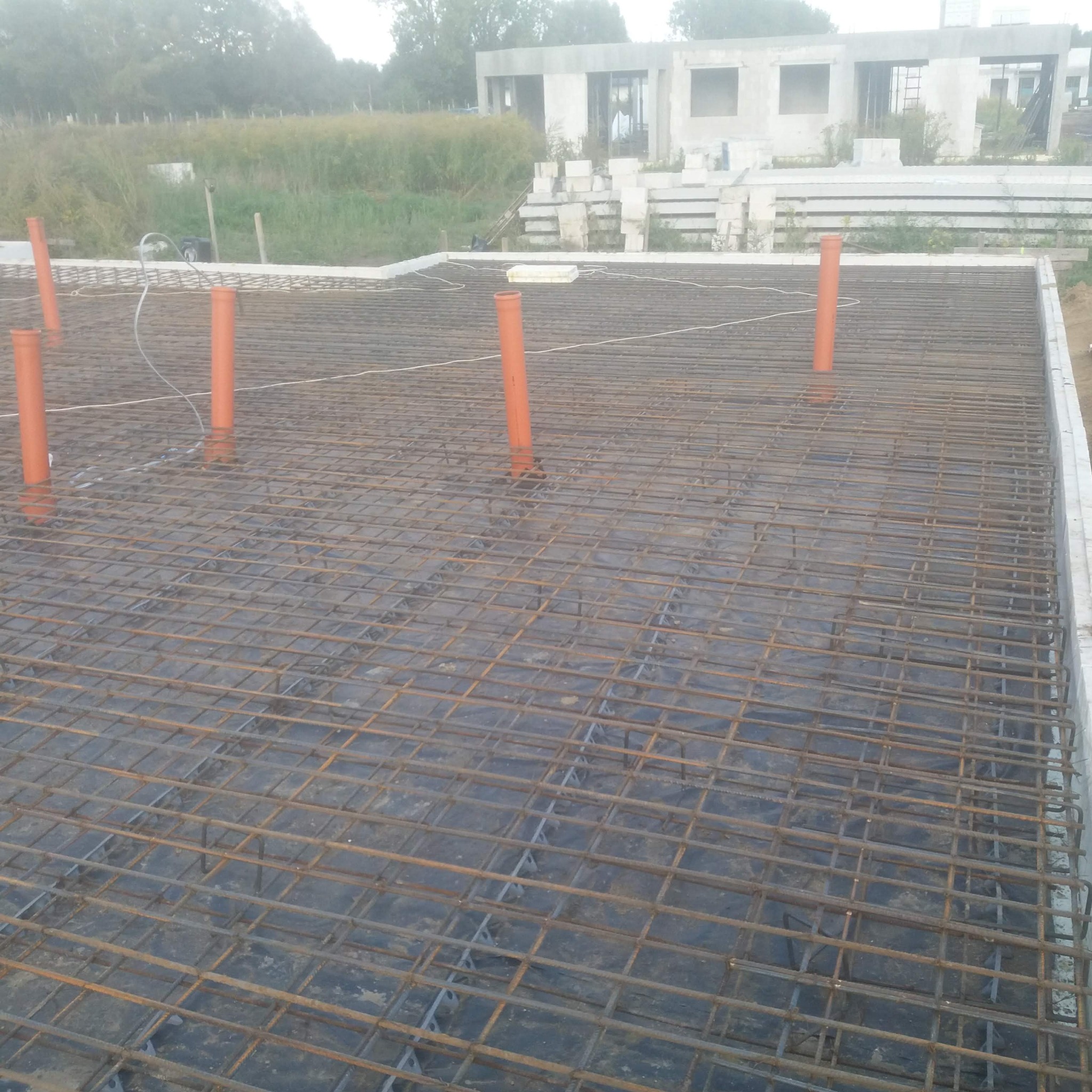 Zbrojenie płyty fundamentowej z wystającymi rurami instalacyjnymi na tle surowego budynku w budowie, widok z góry pod kątem.