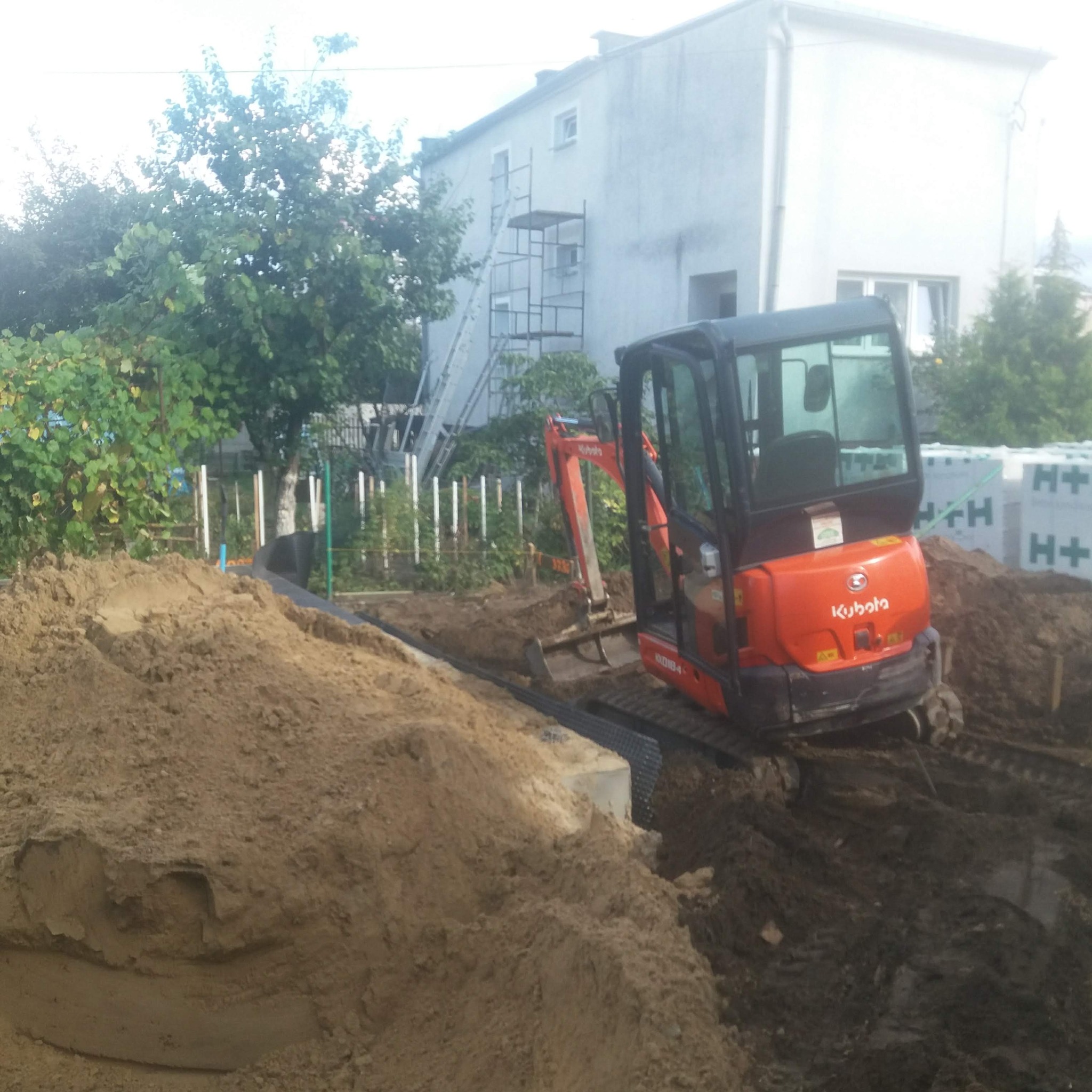 Pomarańczowa minikoparka Kubota na budowie, obok sterta piasku i bloczki H+. Wykop pod fundament zabezpieczony folią. W tle dom i rusztowanie.