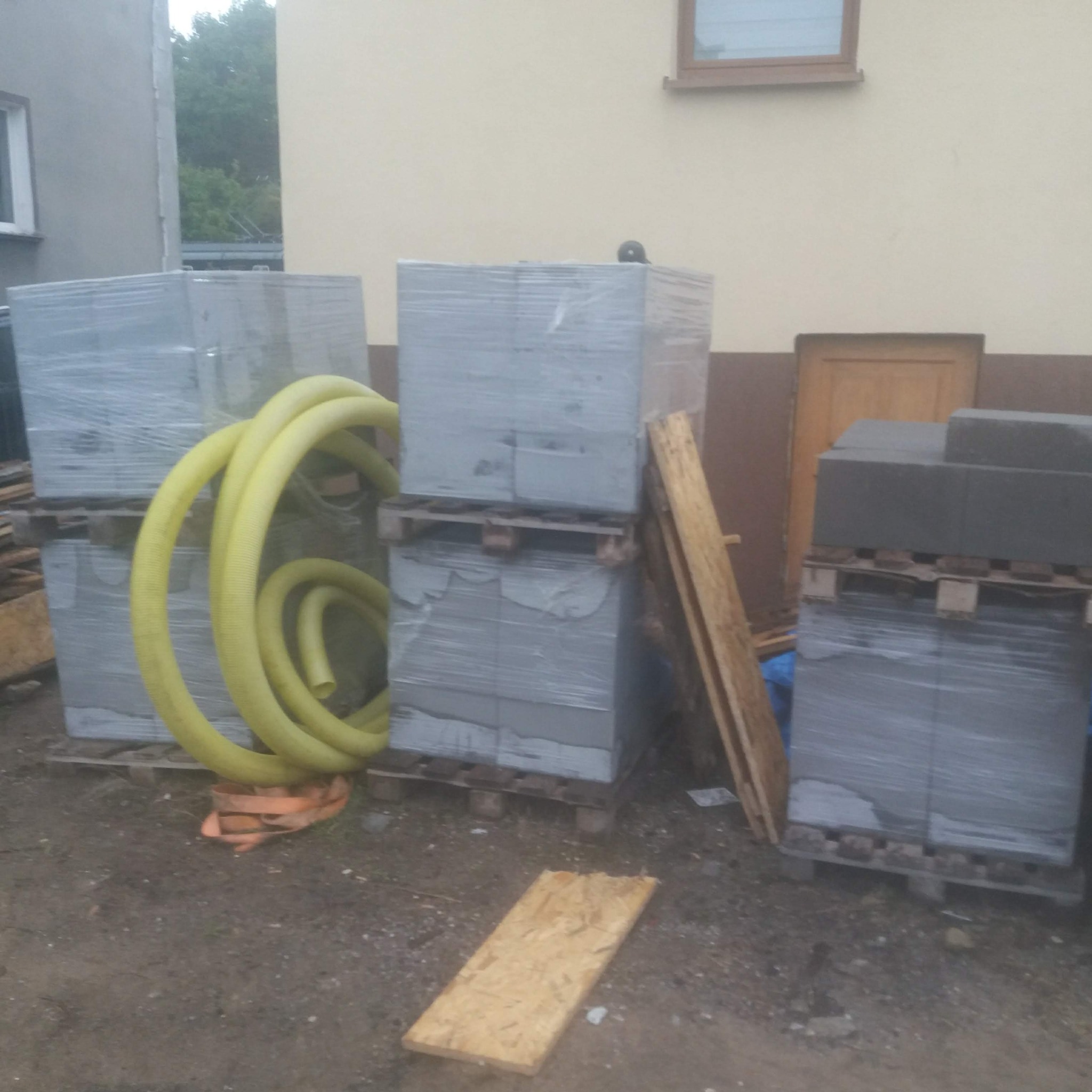 Przygotowane materiały budowlane: palety z bloczkami, żółty wąż wentylacyjny i deski, składowane przy ścianie budynku z oknem. Widok z poziomu gruntu.