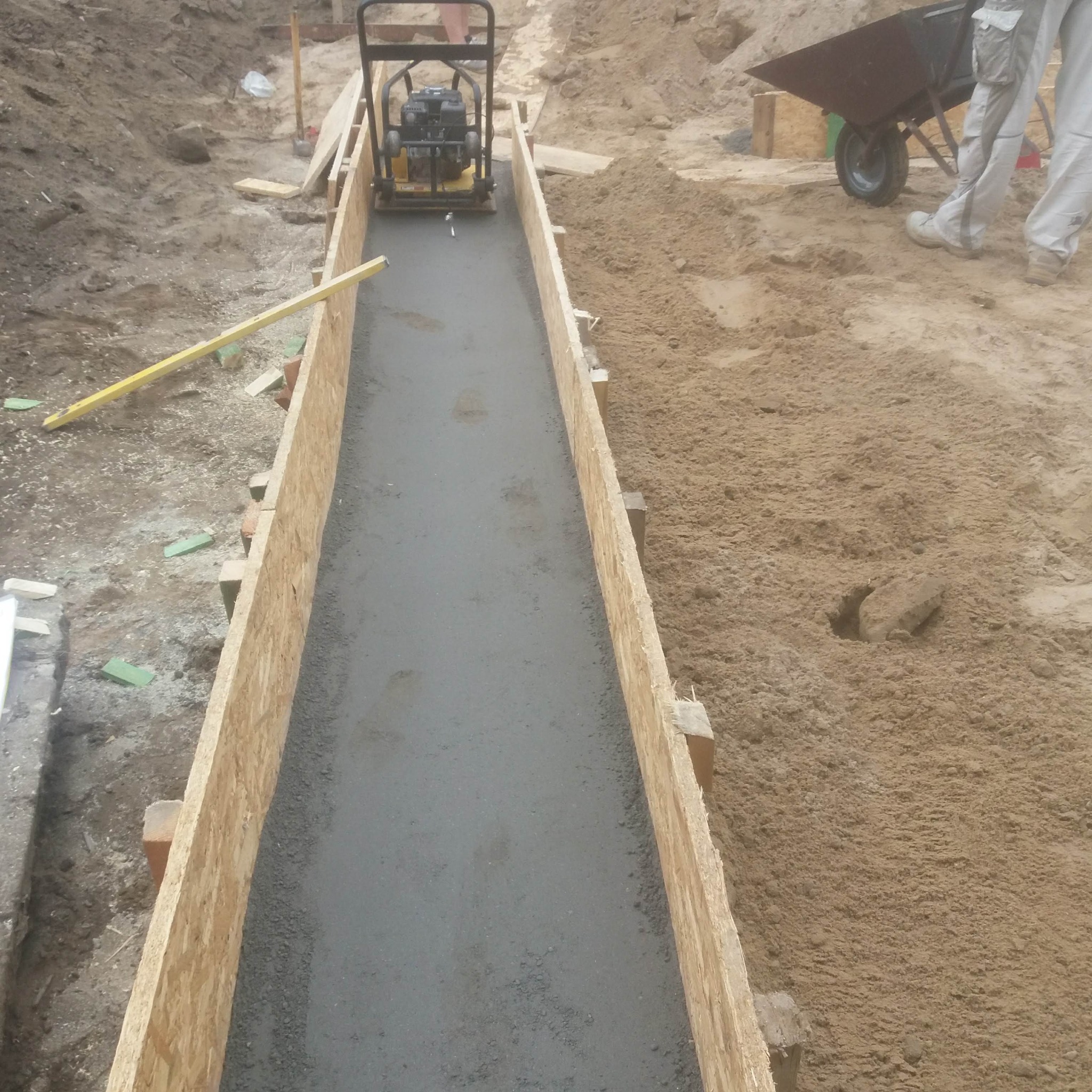 Wyrównywanie świeżej wylewki ubijarką w drewnianym szalunku. W tle piasek, taczka i fragment osoby. Widoczny poziomica.