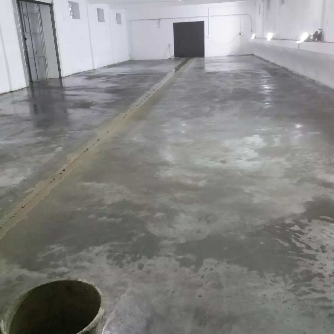 Świeżo wylana, błyszcząca podłoga w hali magazynowej, widoczny betonowy kubeł w rogu. Białe ściany i oświetlenie sufitowe. Ciemne drzwi w tle.