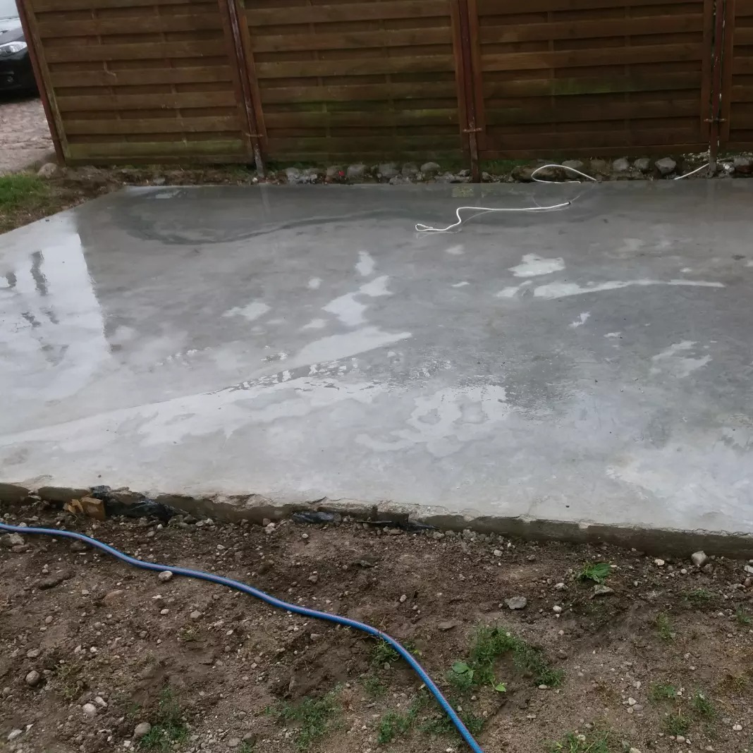 Świeżo wylana, wilgotna powierzchnia w Gorzowie Wielkopolskim. Szara, betonowa płaszczyzna z widocznym wężem i drewnianym ogrodzeniem w tle.