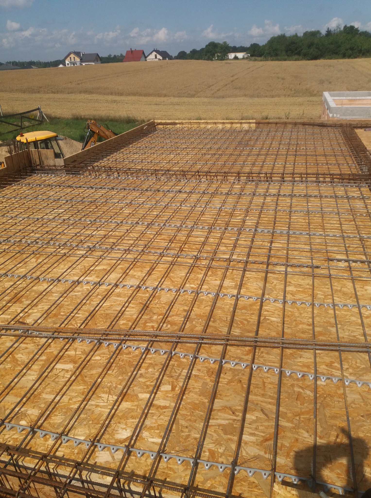 Siatka zbrojeniowa na drewnianym szalunku, przygotowana do zalania betonem. W tle krajobraz polny i zabudowania. Widoczny fragment koparki.
