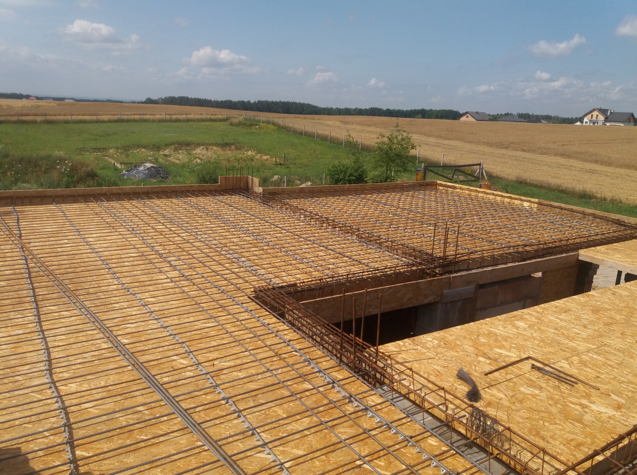Zbrojenie płyty fundamentowej na deskowaniu z płyty OSB, widok z góry na rozległe pole i zabudowania w tle. Przygotowanie do wylewki.