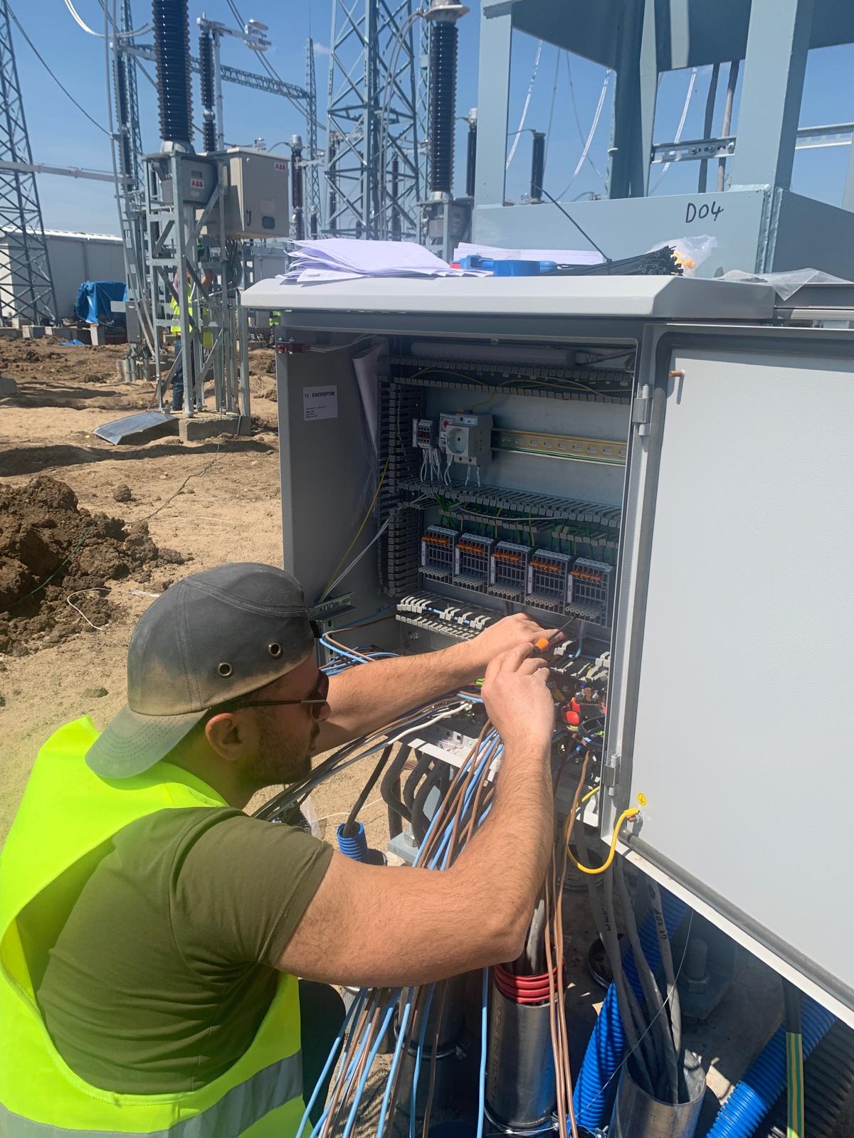 Elektryk w kamizelce montuje przewody w otwartej szafie sterowniczej na tle konstrukcji energetycznej. Przewody o różnych kolorach, narzędzia w dłoniach.