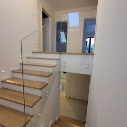 Z-Glass - Nowoczesne schody z drewnianymi stopniami i szklaną balustradą, oświetlone punktowo, prowadzące na wyższą kondygnację w jasnym wnętrzu.