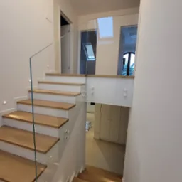 Nowoczesne schody z drewnianymi stopniami i szklaną balustradą, oświetlone punktowo, prowadzące na wyższą kondygnację w jasnym wnętrzu.