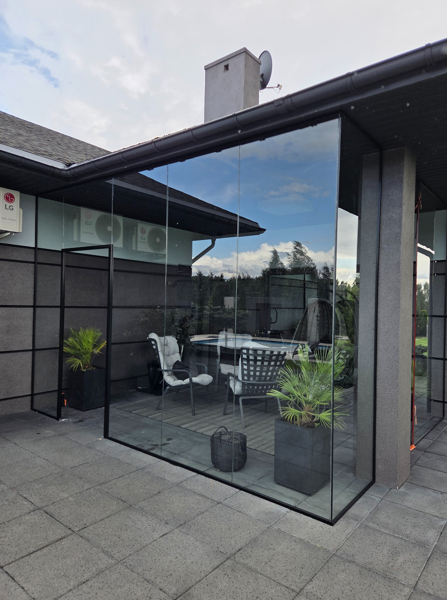 Szklana zabudowa tarasu z czarnymi ramami, widok na meble ogrodowe i rośliny w donicach. Nowoczesny design, szklane ściany odbijają niebo i drzewa.