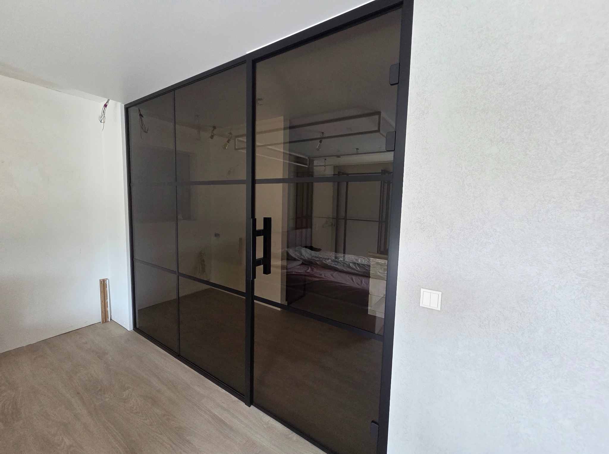 Nowoczesne, przesuwne drzwi szklane z czarną ramą, dzielone na prostokątne segmenty. Widok na wnętrze pokoju z łóżkiem. Minimalistyczny design.