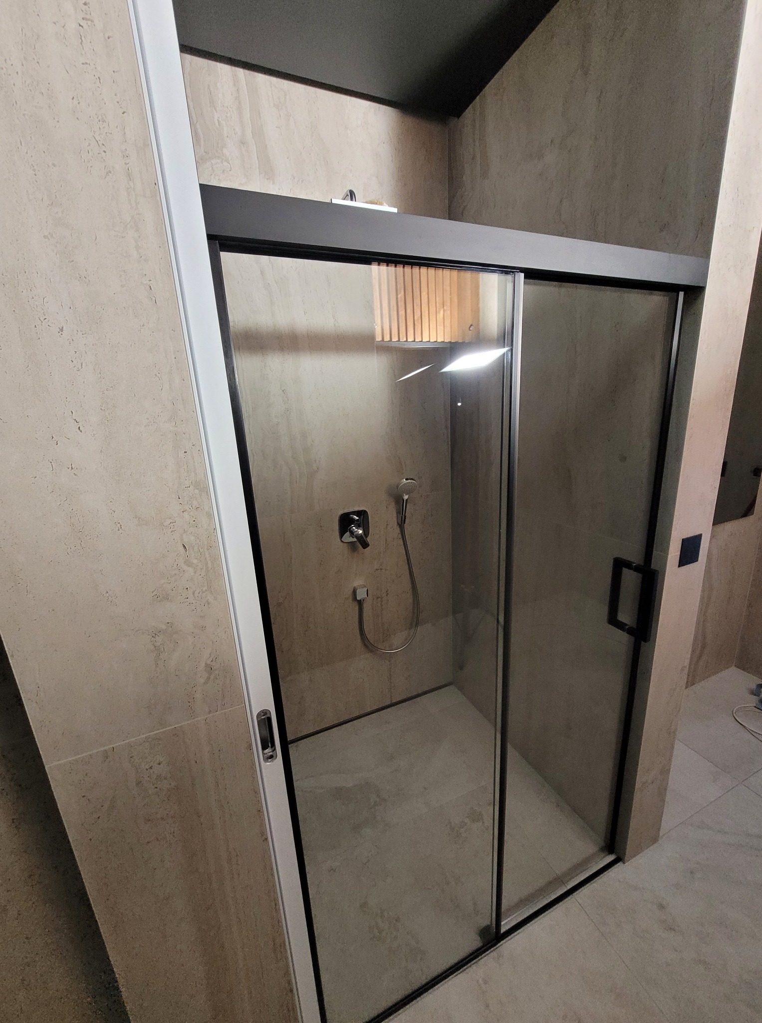 Nowoczesna kabina prysznicowa z przesuwnymi drzwiami szklanymi, czarne profile, jasne płytki imitujące kamień, minimalistyczna armatura.