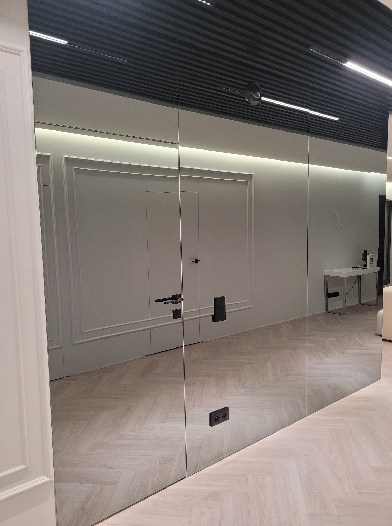 Szklane drzwi z czarną klamką, odbijające wzór parkietu i fragment wnętrza. Minimalistyczny design, widoczne łączenia tafli szkła. Eleganckie wykończenie w nowoczesnym stylu.