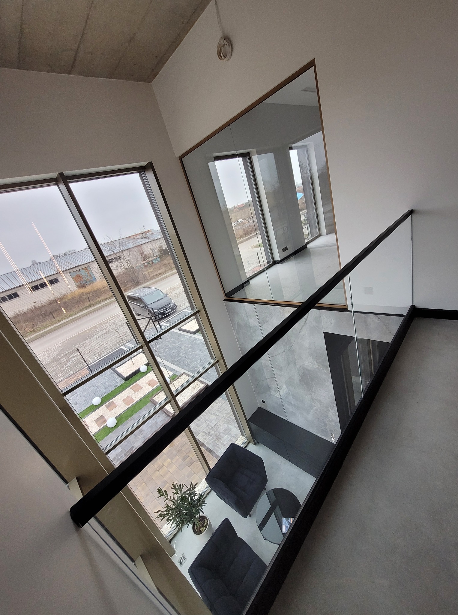 Wnętrze domu z nowoczesną szklaną balustradą schodową z czarnymi elementami. Widok z góry na salon z szarymi fotelami i stolikiem. Duże okna z widokiem na ulicę.