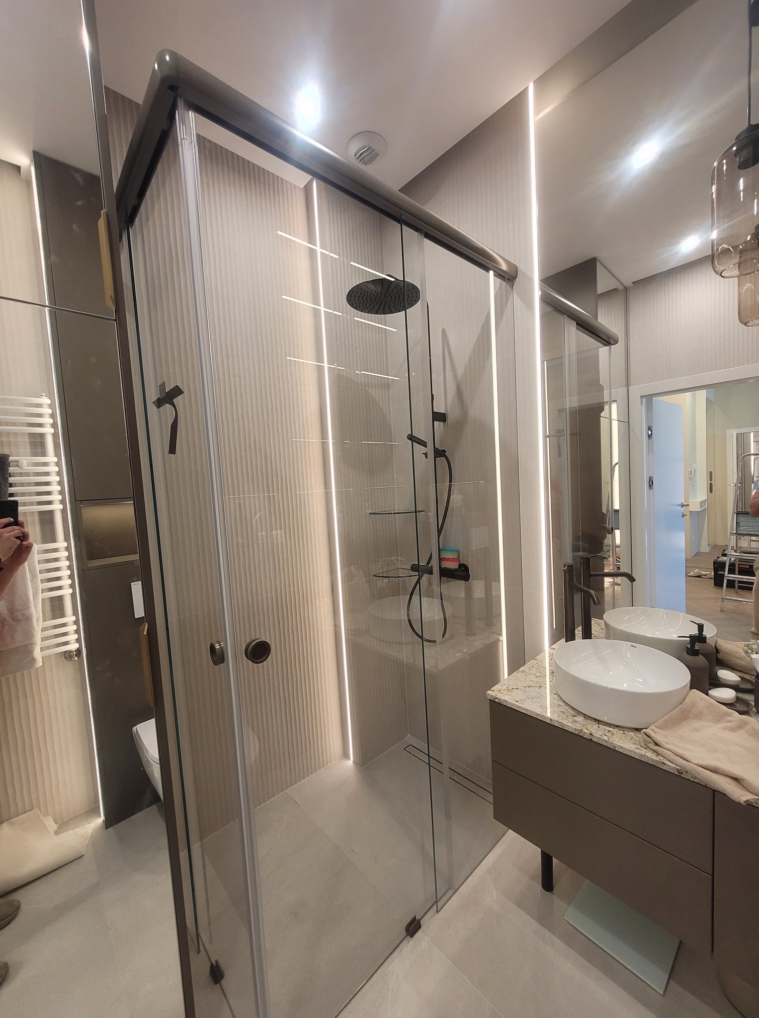 Nowoczesna kabina prysznicowa ze szklanymi drzwiami, oświetleniem LED i elegancką armaturą w łazience. Widoczne umywalka i lustro.