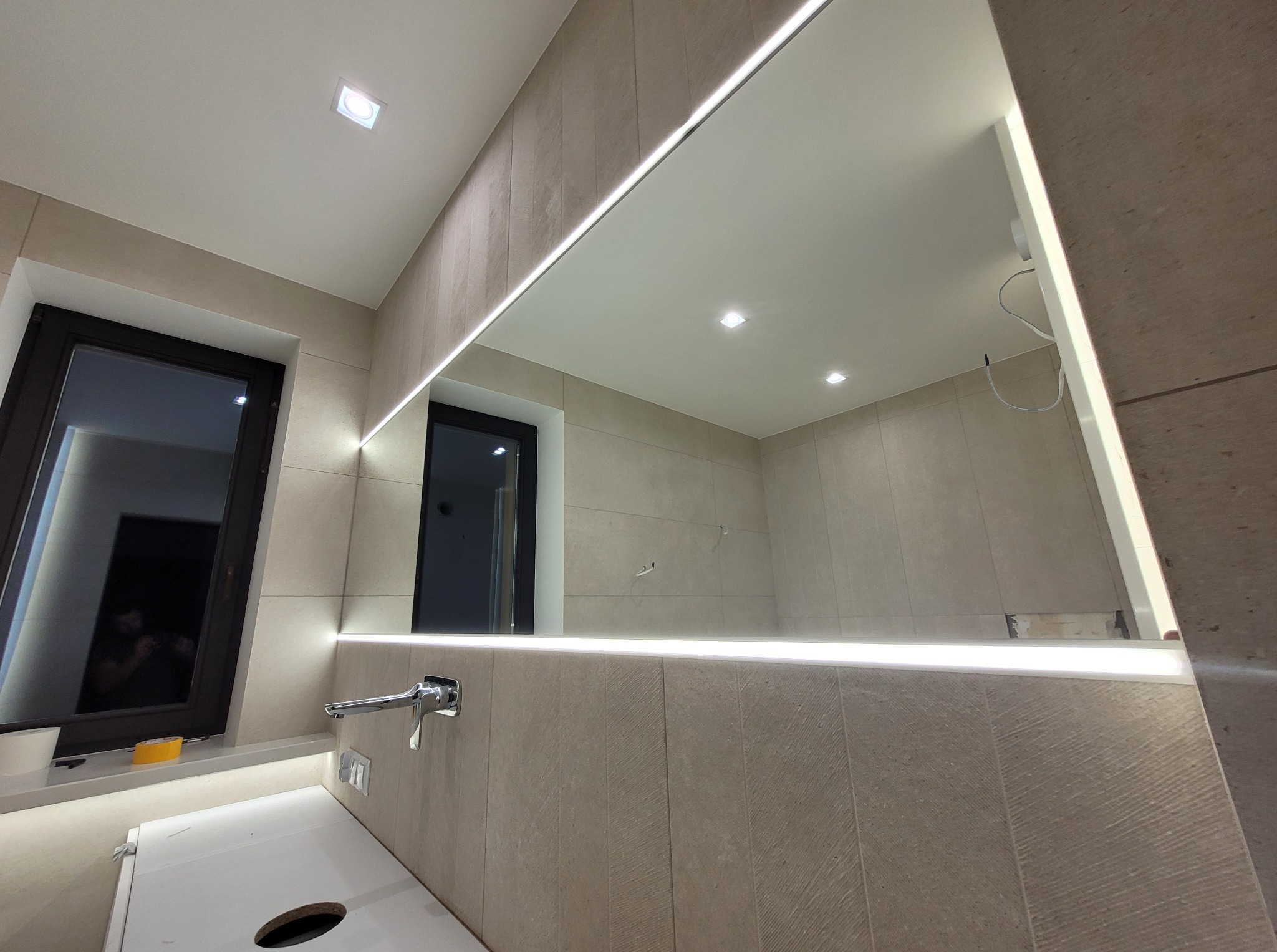 Nowoczesna łazienka z podświetlanym lustrem i armaturą. Widoczne okno i elementy wykończenia w stonowanej kolorystyce. Minimalistyczny design z dbałością o detale.