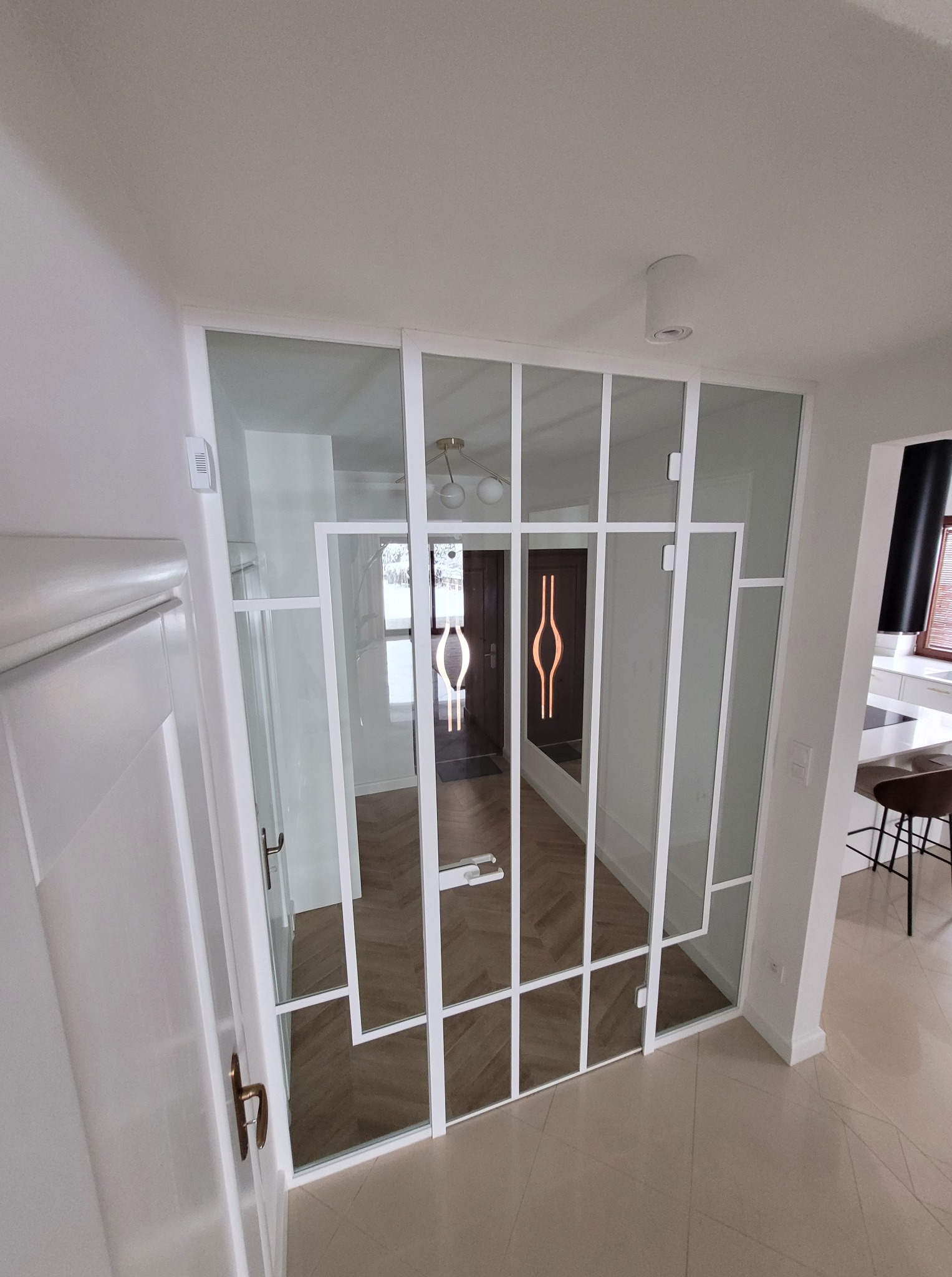 Nowoczesna, szklana ścianka działowa z białymi profilami, oddzielająca korytarz od reszty domu. Widoczny parkiet i elementy wystroju wnętrza.