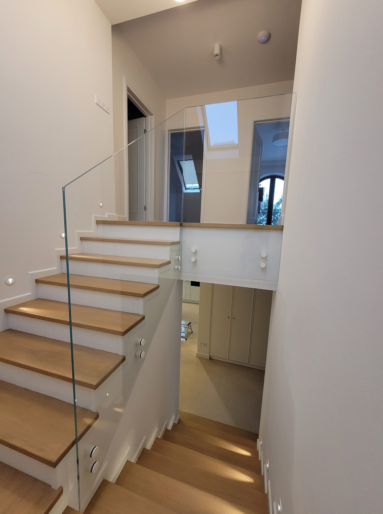 Nowoczesne schody z drewnianymi stopniami i szklaną balustradą, oświetlone punktowo, prowadzące na wyższą kondygnację w jasnym wnętrzu.