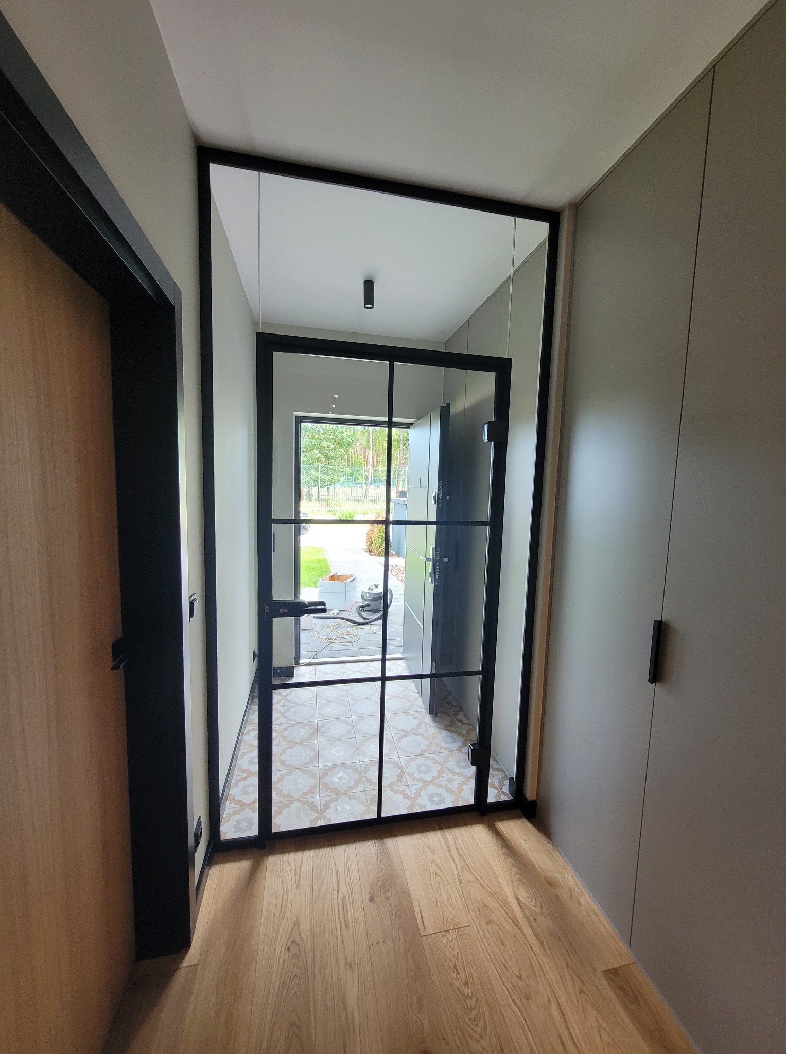 Nowoczesne drzwi szklane z czarną ramą i podziałami, widok z wnętrza domu na ogród. Jasne wnętrze, parkiet i szafa w zabudowie.