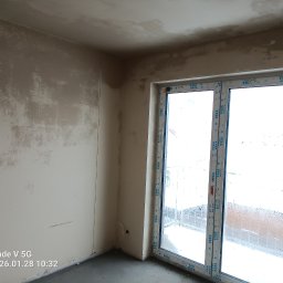STYLBUD KAZIMIERZ BUTKIEWICZ - Pokój w trakcie prac tynkarskich: ściany pokryte warstwą tynku, widoczne okno balkonowe z ramą VEKA. Surowy stan wykończenia wnętrza.