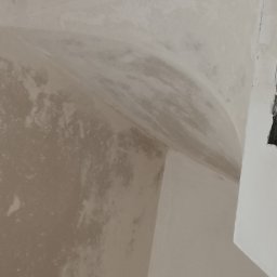 STYLBUD KAZIMIERZ BUTKIEWICZ - Niedokończone tynki na suficie i ścianie, widoczne nierówności i zacieki. Fragment okna w prawym górnym rogu. Ujęcie z dołu, blisko powierzchni.