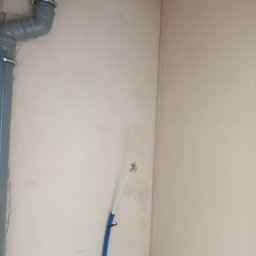 STYLBUD KAZIMIERZ BUTKIEWICZ - Ściana po tynkowaniu maszynowym z widoczną instalacją hydrauliczną i rurą kanalizacyjną. Jasne, neutralne tło. Widoczne detale instalacji.