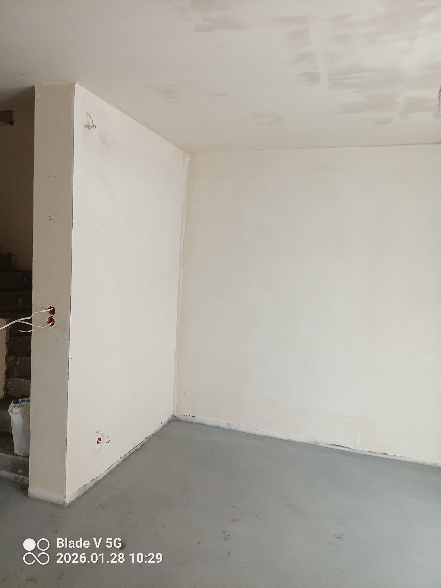 Świeżo otynkowane, gładkie ściany w rogu pomieszczenia z widocznymi puszkami elektrycznymi i wylewką podłogową. Widoczne schody w tle.