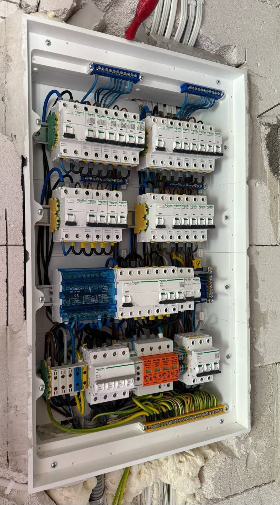 Rozdzielnica elektryczna Schneider w białej obudowie, z licznymi zabezpieczeniami i okablowaniem wewnątrz surowego muru. Widoczne złącza, bezpieczniki i przewody.