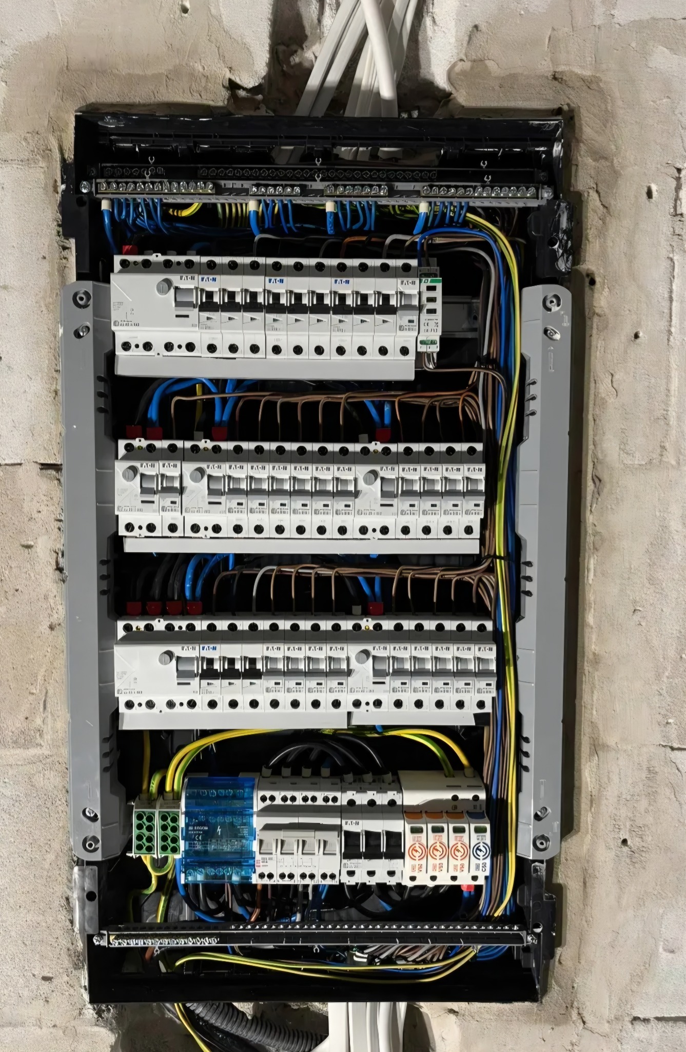 Rozdzielnica elektryczna z bezpiecznikami i przewodami w szarej obudowie, zamontowana na surowej ścianie z betonu komórkowego. Widoczne kable w różnych kolorach.