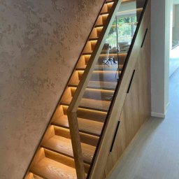 Luxhome Remonty - Nowoczesne schody drewniane z podświetleniem LED i szklaną balustradą, zintegrowane z szafą wnękową, na tle ściany o dekoracyjnej fakturze.