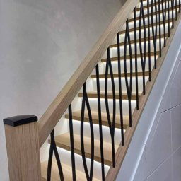 Luxhome Remonty - Nowoczesne schody z drewnianymi stopniami, podświetleniem LED i czarną, dekoracyjną balustradą. Jasne drewno i minimalistyczny design. Ujęcie z dołu.