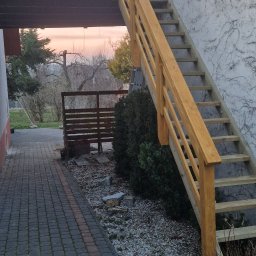 Luxhome Remonty - Zewnętrzne, drewniane schody z balustradą, prowadzące na piętro budynku w Ustrzykach Dolnych. Widok na chodnik z kostki i krzewy.