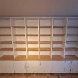 Luxhome Remonty - Biała zabudowa meblowa z drewnianymi półkami i szafkami z folią ochronną. Widok na regał wnękowy z otwartymi półkami i dolnymi szafkami.