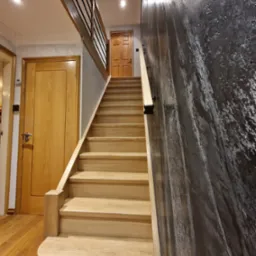 Jasne, drewniane schody z poręczą, prowadzące do g&oacute;ry, kontrastujące z ciemną, teksturowaną ścianą. Widoczne drzwi na g&oacute;rze i dole schod&oacute;w.