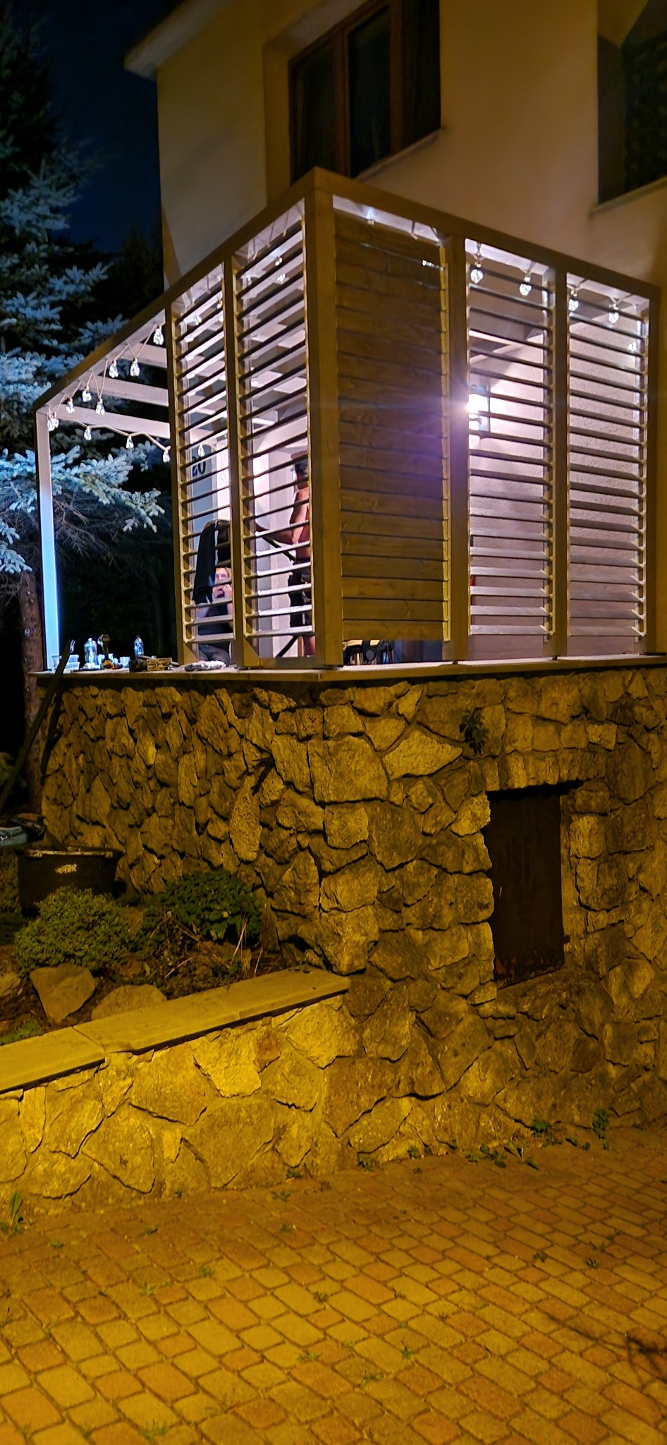 Oświetlona, drewniana konstrukcja tarasowa z żaluzjami, wzniesiona na kamiennej podstawie. Widoczne oświetlenie i elementy dekoracyjne.