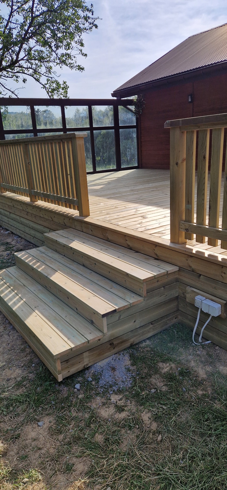 Jasne drewniane schody prowadzą na taras z balustradami i przeszkloną ścianą. Widoczna elewacja drewnianego budynku i zieleń w tle.