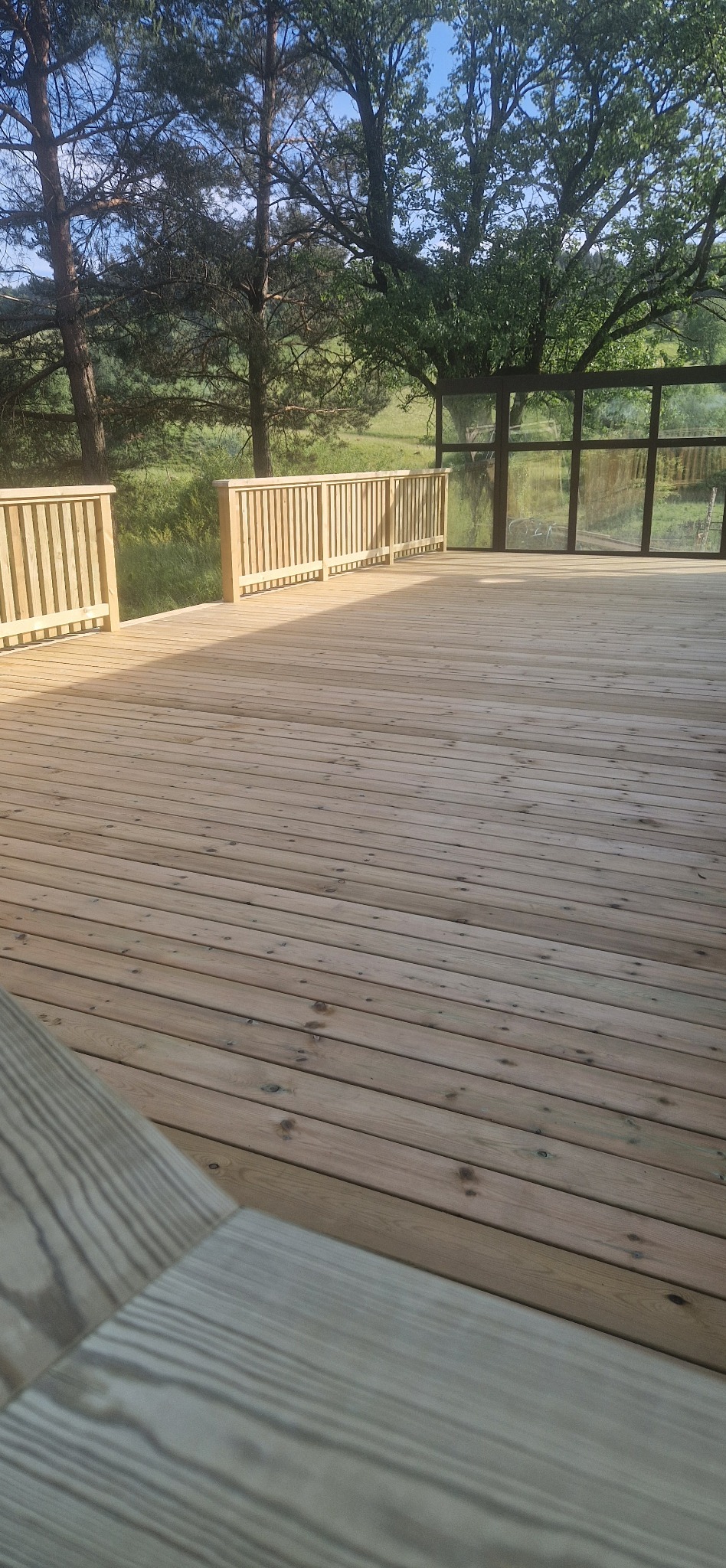 Jasny taras z desek z drewnianą balustradą i widokiem na zieleń. Ujęcie w słoneczny dzień, z widocznymi detalami tekstury drewna i otaczającej przyrody.