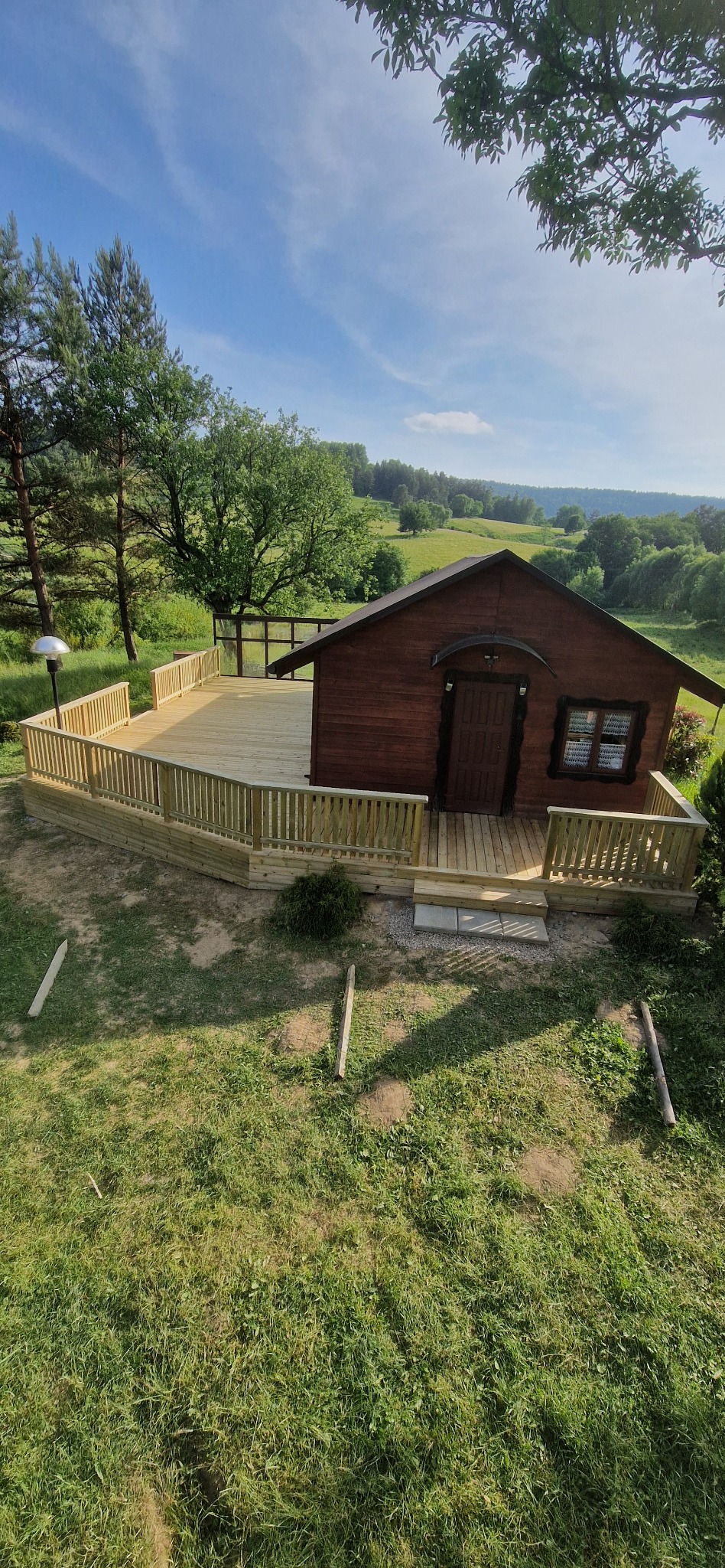 Drewniany taras z balustradą otaczający domek na wzgórzu, widok z góry. Jasne deski tarasu kontrastują z ciemnym kolorem domku i zielenią trawy.