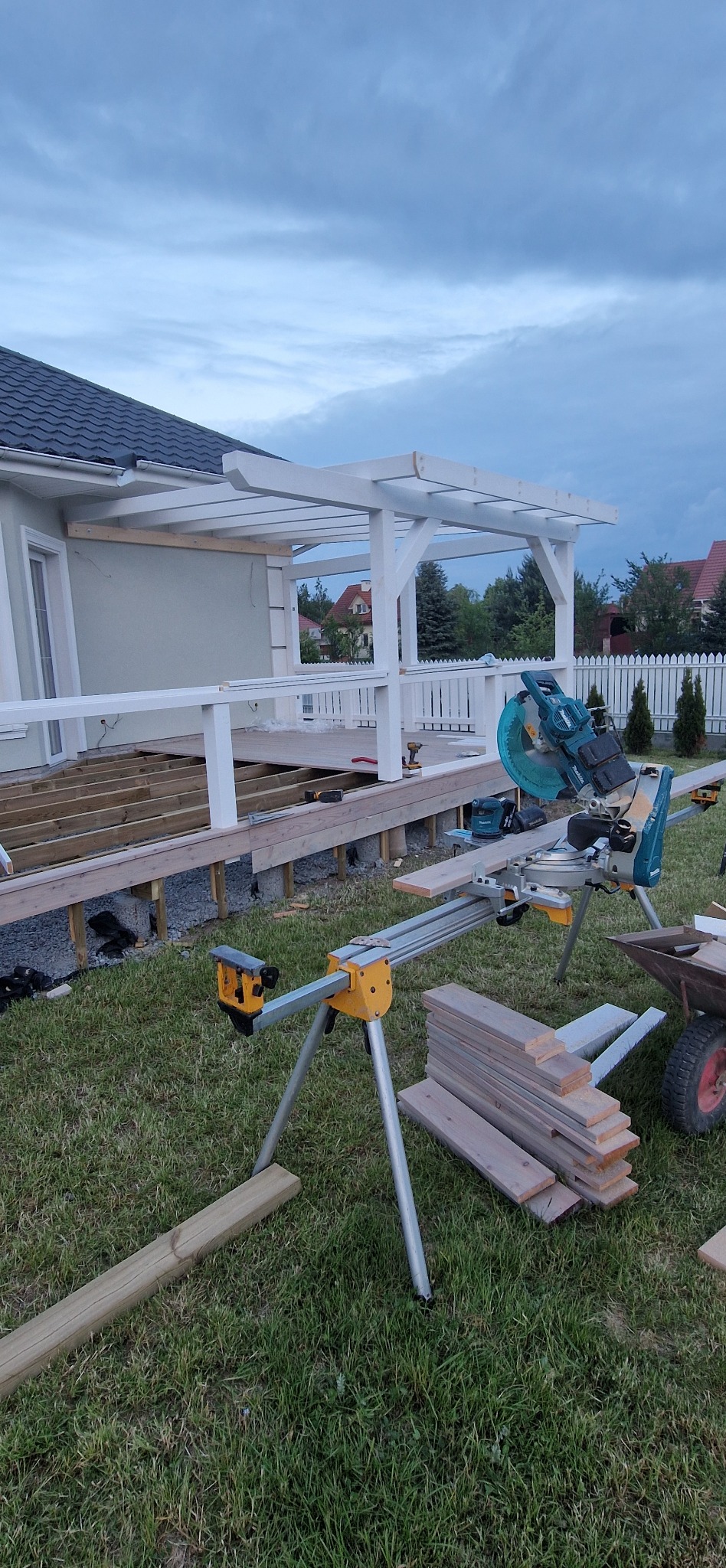 Budowa tarasu z białą pergolą w ogrodzie. Widoczna piła do drewna, deski i elementy konstrukcyjne. Prace w toku, widok na dom i ogrodzenie.