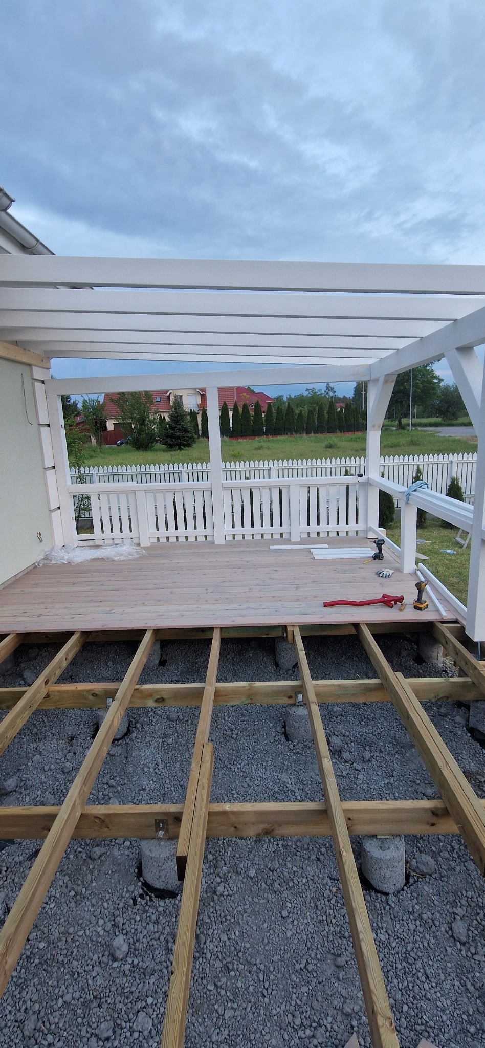 Montaż tarasu: drewniana konstrukcja na betonowych podporach, częściowo ułożone deski, narzędzia widoczne na jasnym tle. W tle białe ogrodzenie.