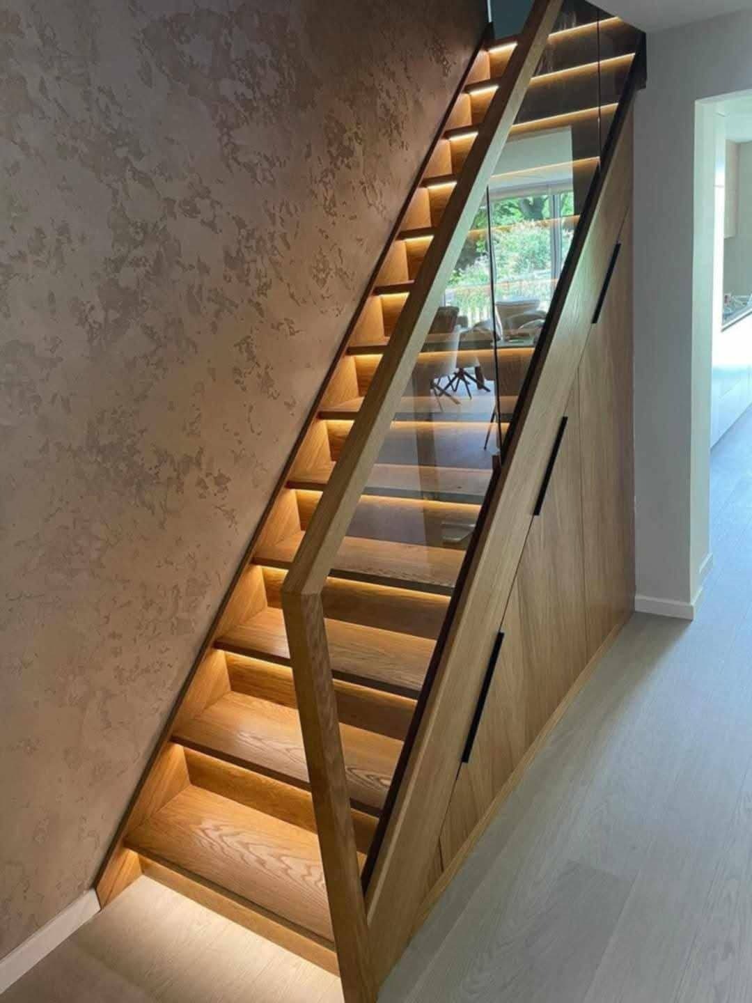 Nowoczesne schody drewniane z podświetleniem LED i szklaną balustradą, zintegrowane z szafą wnękową, na tle ściany o dekoracyjnej fakturze.