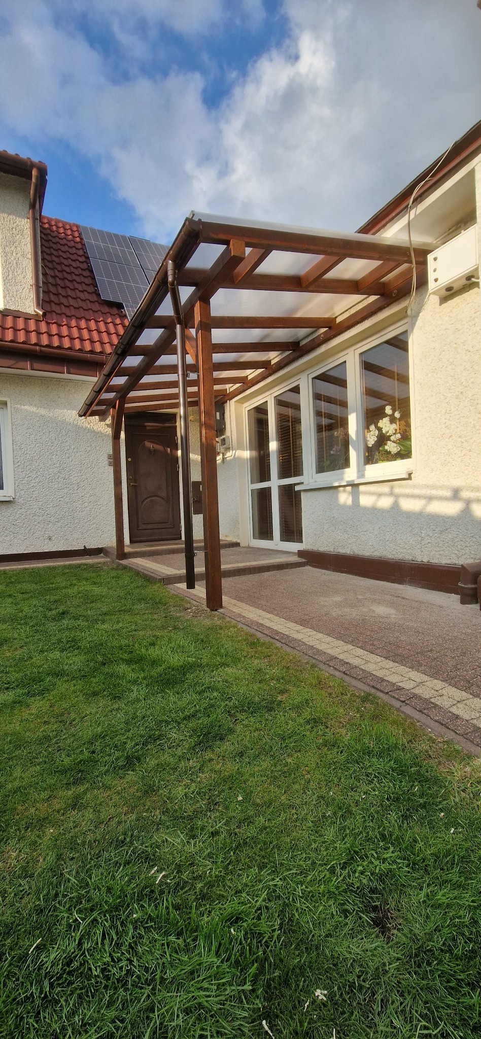 Zadaszenie wejścia do domu z drewnianą konstrukcją i przezroczystym dachem, widoczne panele słoneczne na dachu domu, trawnik i fragment chodnika.