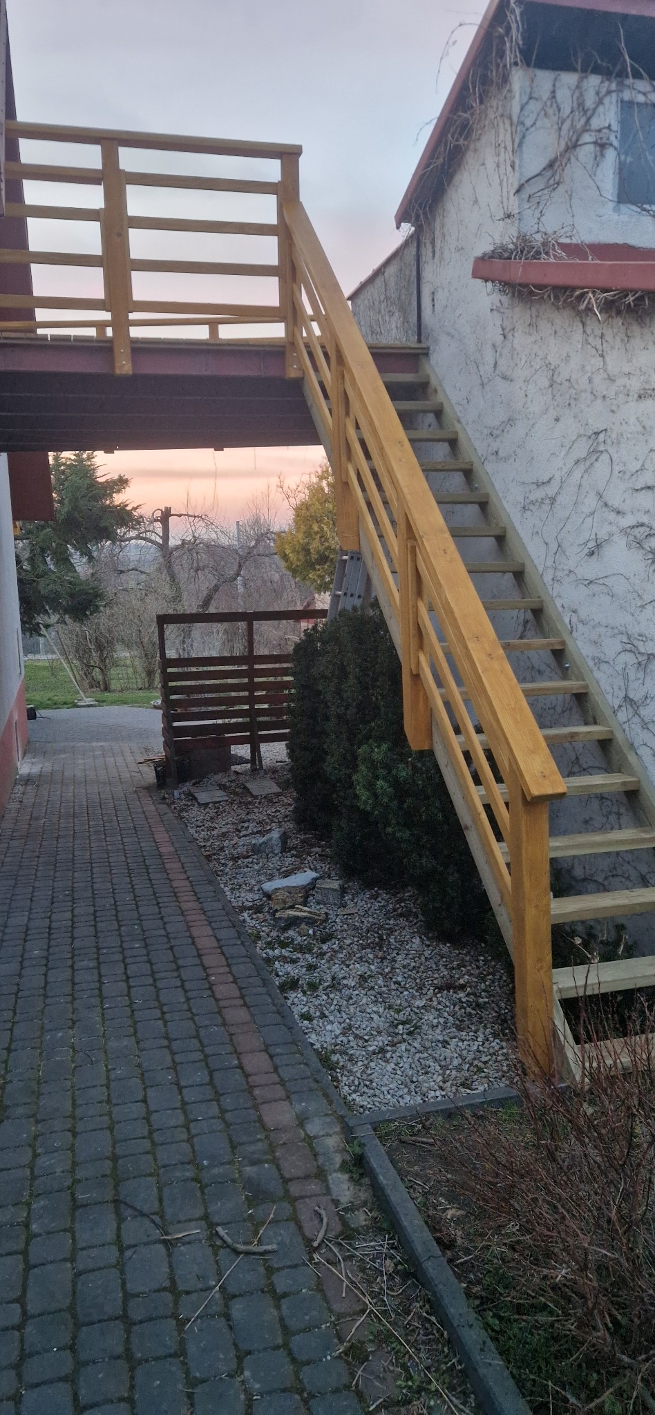 Zewnętrzne, drewniane schody z balustradą, prowadzące na piętro budynku w Ustrzykach Dolnych. Widok na chodnik z kostki i krzewy.